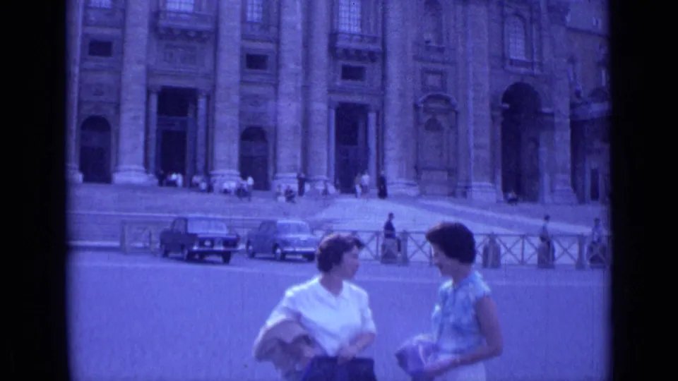 The women continue talking in a 1962 Rome street view, framed against the basilica entrance, steps, railings, and period cars.