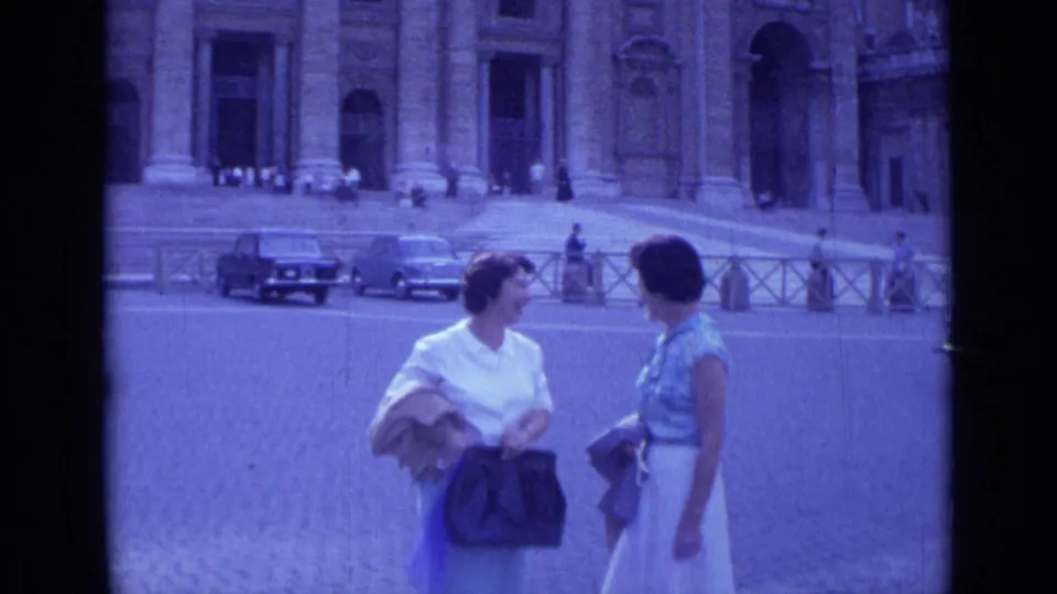 A closer 1962 Rome home-movie frame shows the two women talking near the camera with St. Peter's Basilica and mid-century cars behind.