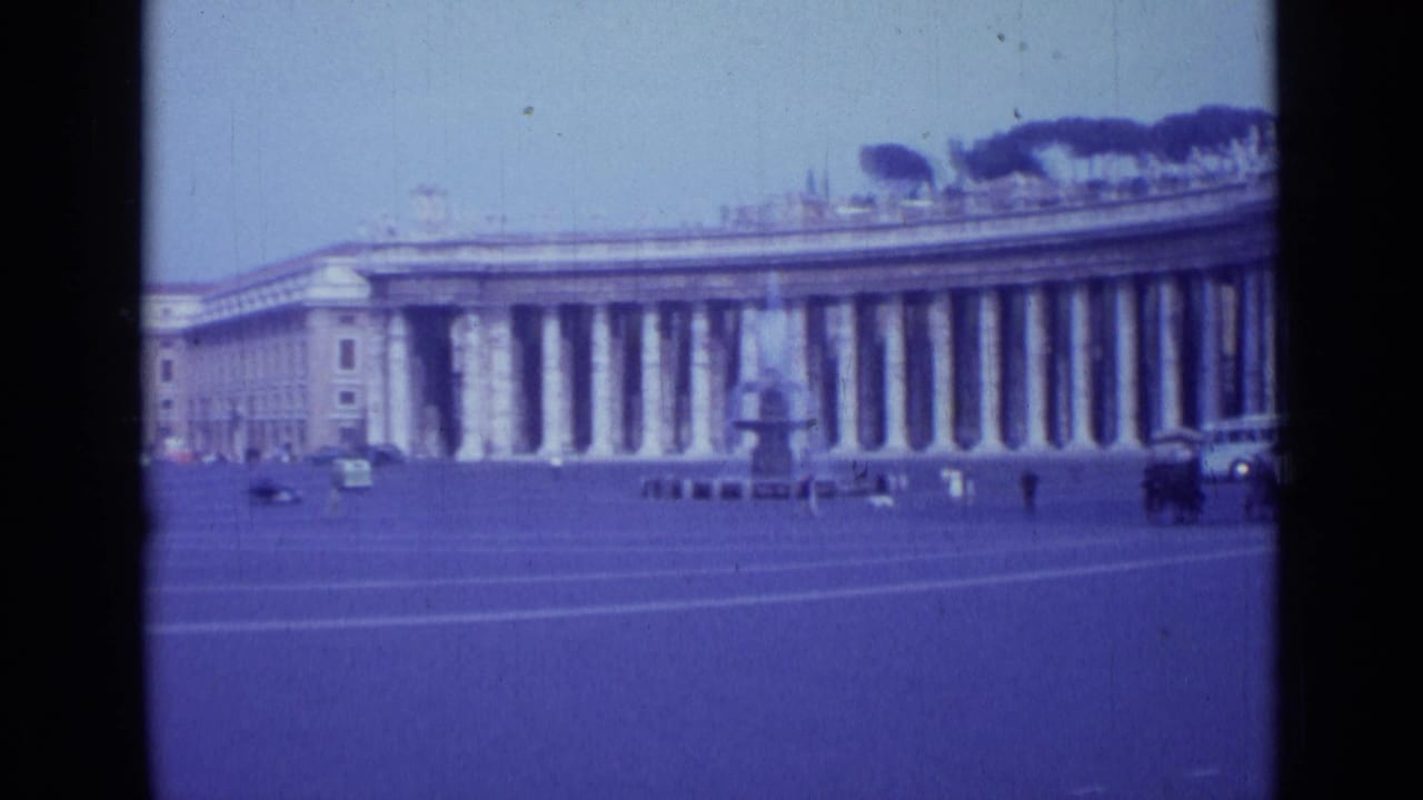 Wide square view with colonnade, fountain, cars, and pedestrians in the distance.