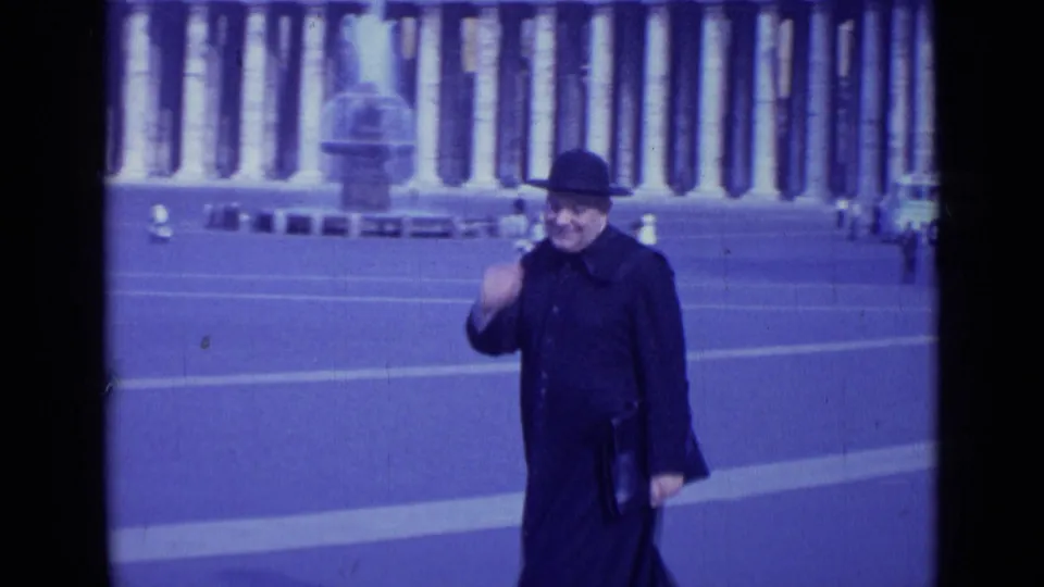 A smiling man in a dark hat and coat waves toward the 8mm camera in St. Peter's Square, Rome.