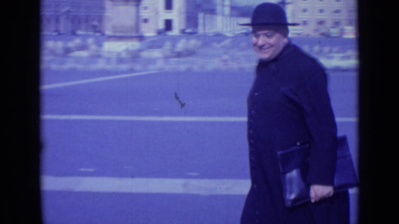 In 1962 Rome, a man in a dark coat and bowler-style hat walks across a broad plaza while looking toward the camera.