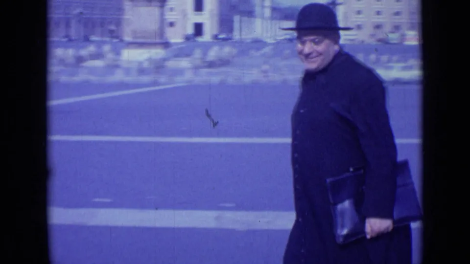 In 1962 Rome, a man in a dark coat and bowler-style hat walks across a broad plaza while looking toward the camera.