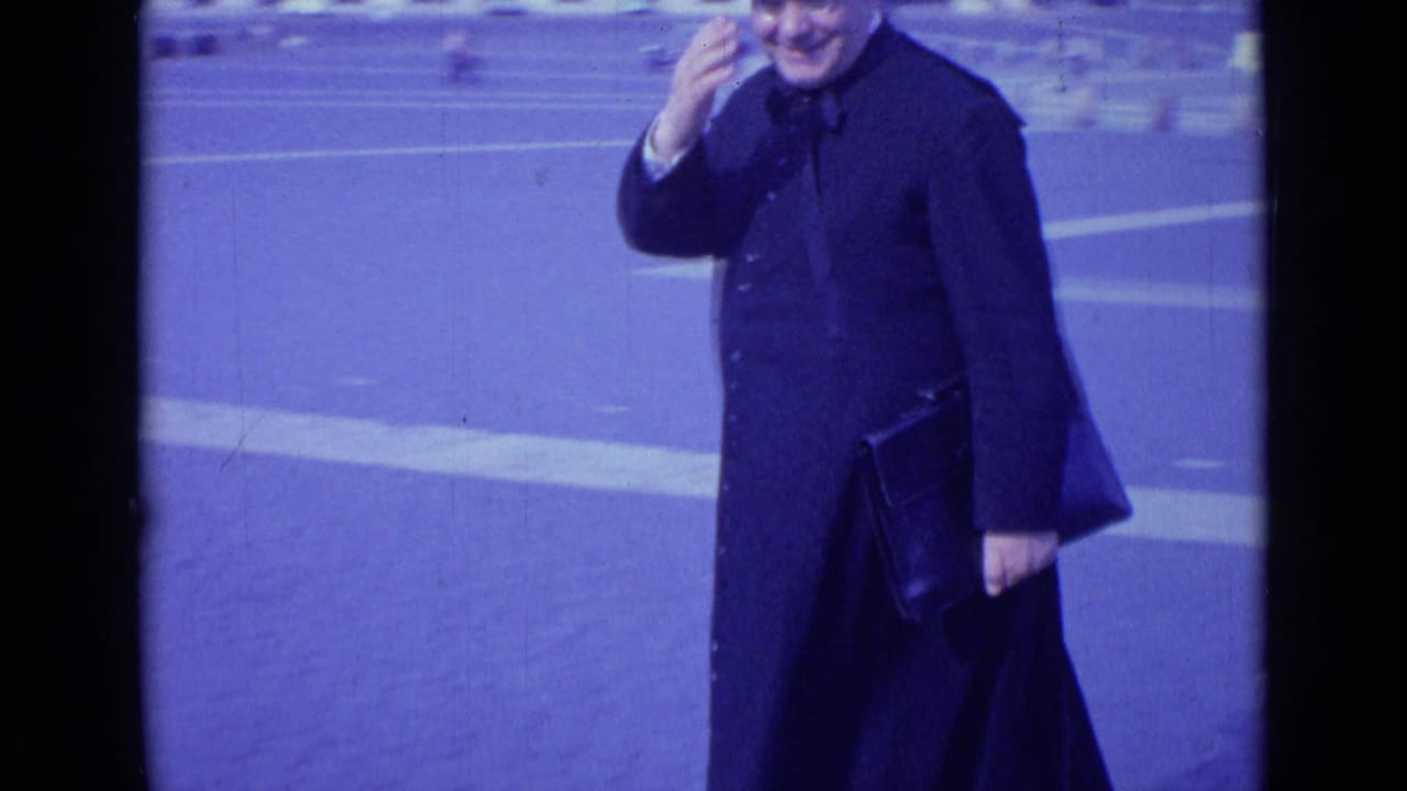 Smiling man in dark coat waving toward the camera.
