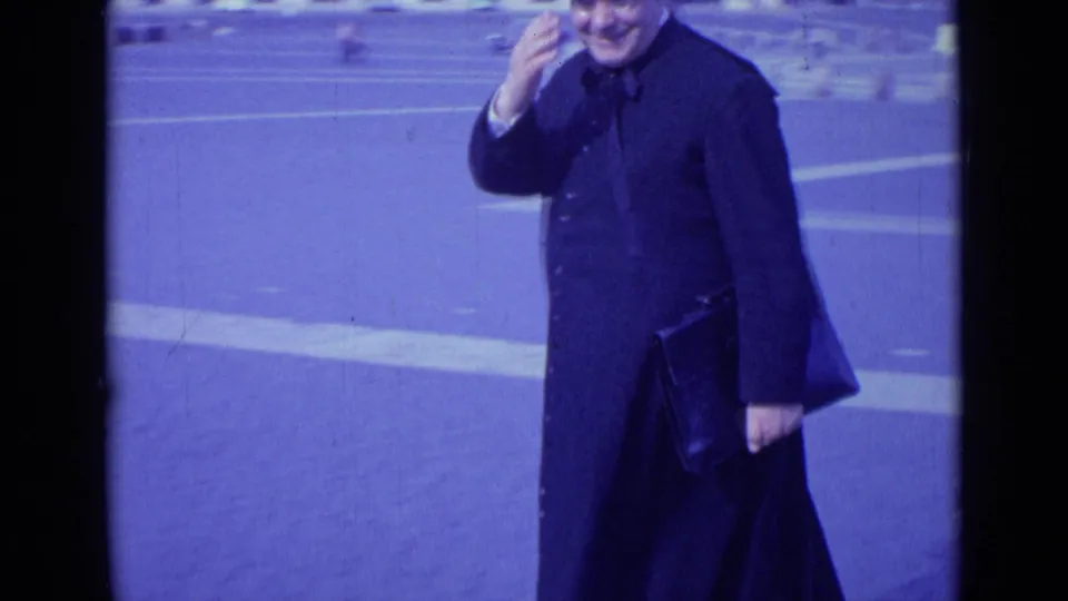 In 1962 Rome, the man smiles and raises a hand toward the camera while holding a dark leather case.