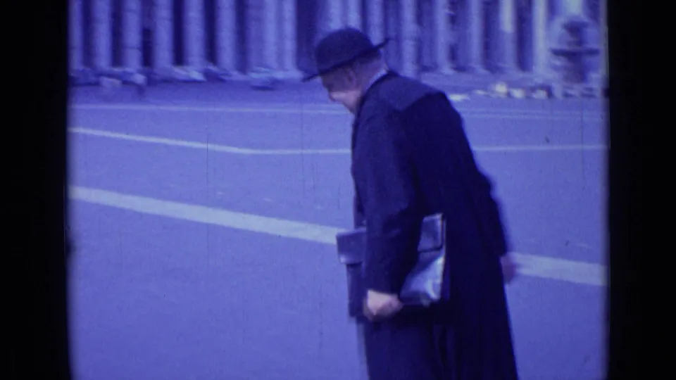 In 1962 Rome, the man turns away from the camera, carrying a leather case across the open paved area.