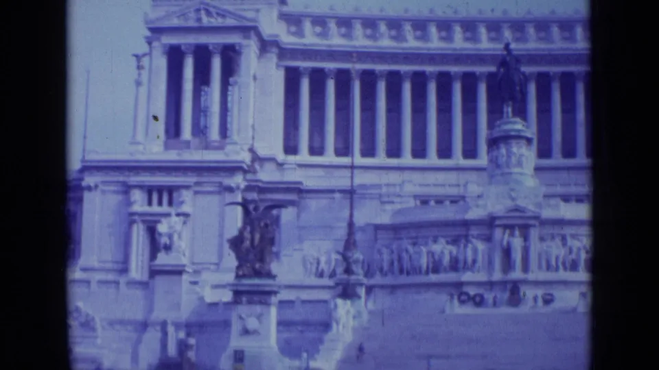 In 1962 Rome, the camera frames a grand public facade with columns, sculptural figures, and ornate stone monuments.