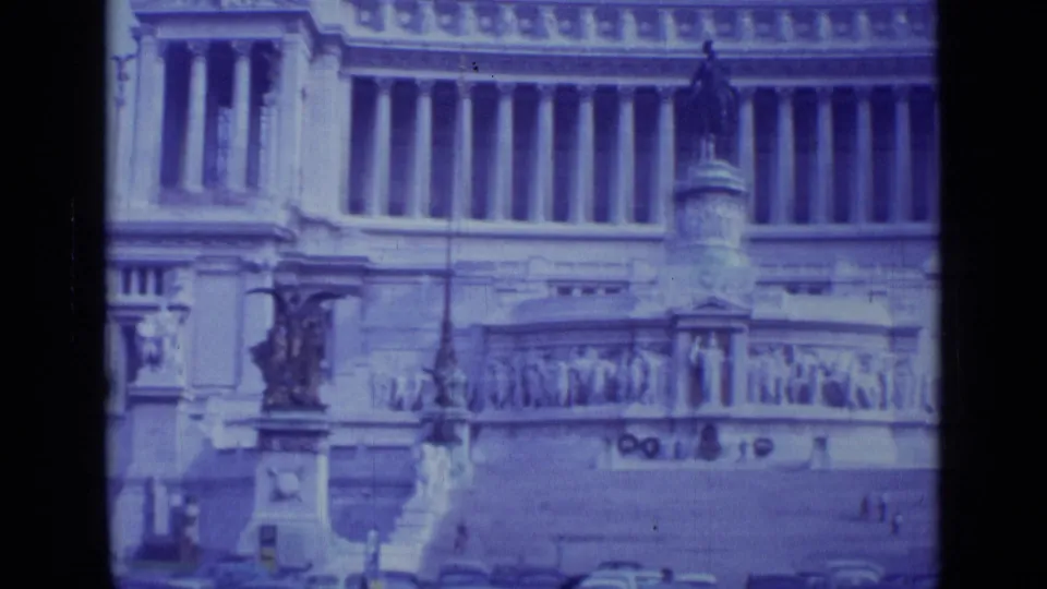 1962 Rome 8mm frame showing a monumental stone facade with columns, relief sculpture, steps, a statue, and tiny figures in the plaza.