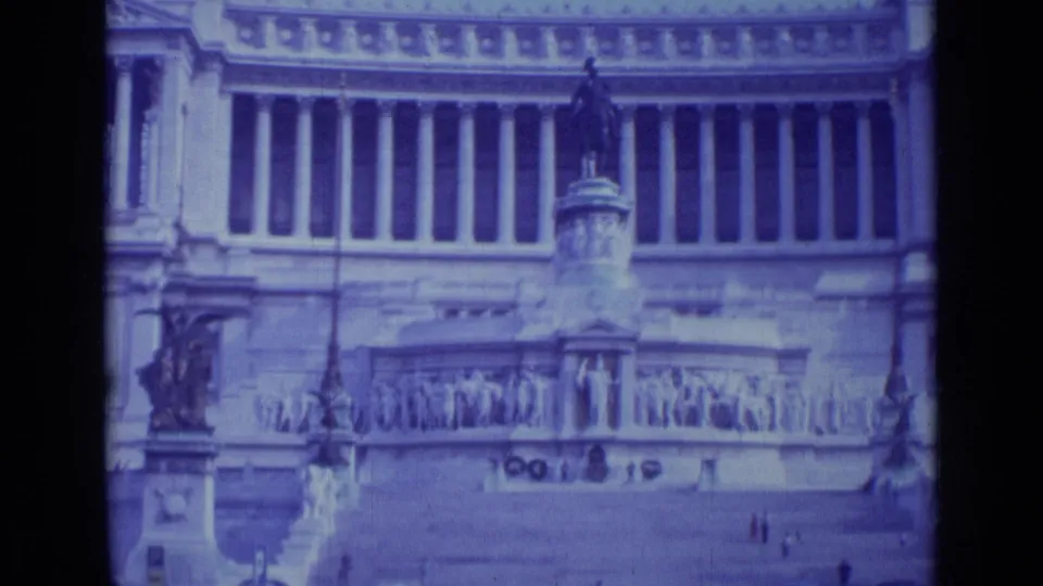 1962 Rome 8mm frame of tall columns, carved reliefs, steps, and a foreground statue seen across an open plaza.