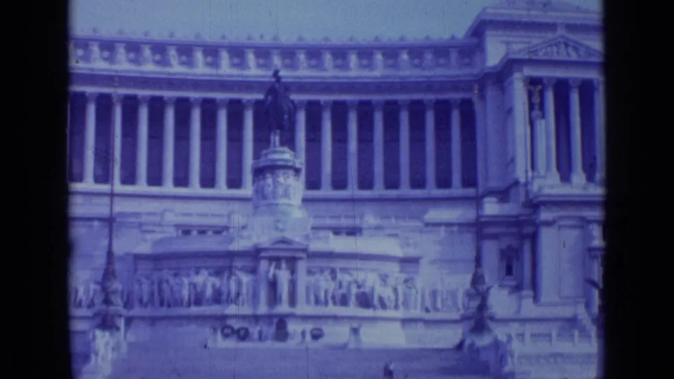 1962 Rome 8mm frame showing a closer visitor view of the monument's columns, statue base, relief carvings, and open plaza.