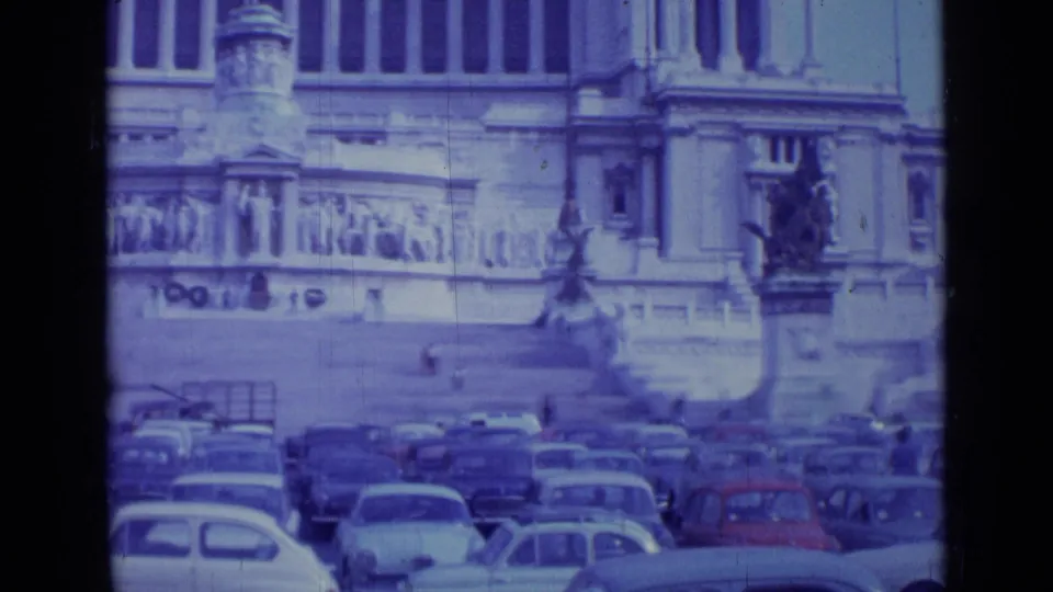 1962 Rome frame with compact cars filling the foreground and monumental stone architecture visible across the square.