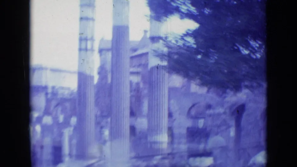 1962 Rome frame showing classical columns, stone remains, and surrounding trees in soft blue-purple 8mm texture.