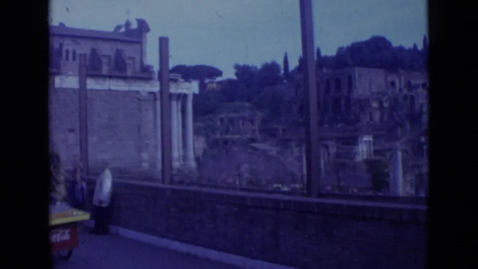 1962 Rome frame with tourists near a brick parapet, classical columns, ruined walls, and umbrella pines in the background.