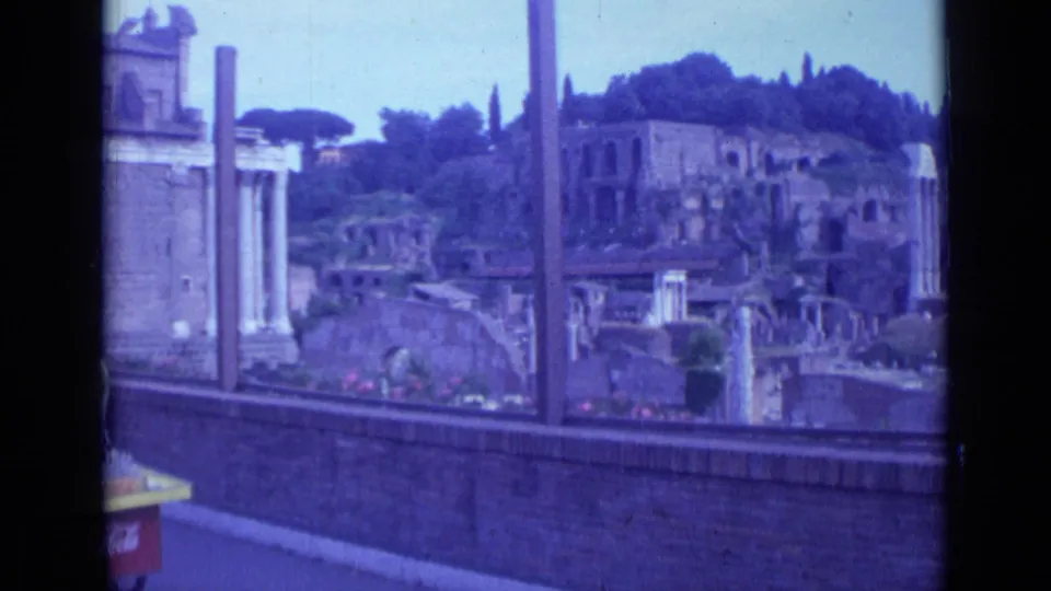 Handheld 1962 Rome view across a brick parapet toward ancient ruins, upright columns, hillside trees, and part of a street cart.