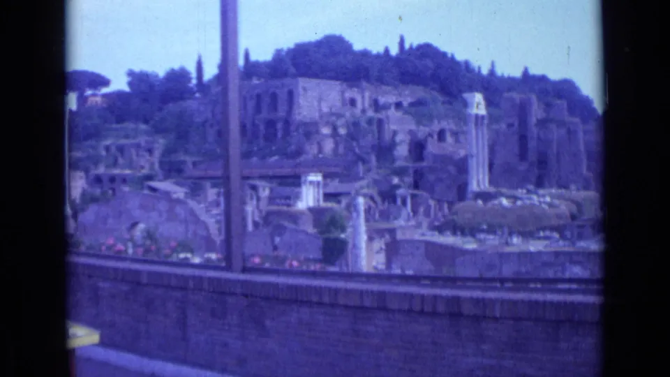 1962 Rome 8mm frame showing ruined stone walls, column fragments, a brick parapet, vertical pole, and dark trees above the site.