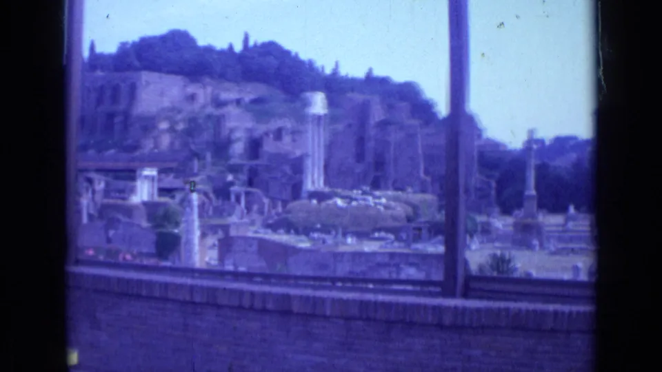 Rome ruins appear behind dark window framing, with columns, weathered masonry, hillside trees, and pale sky visible on 8mm film.