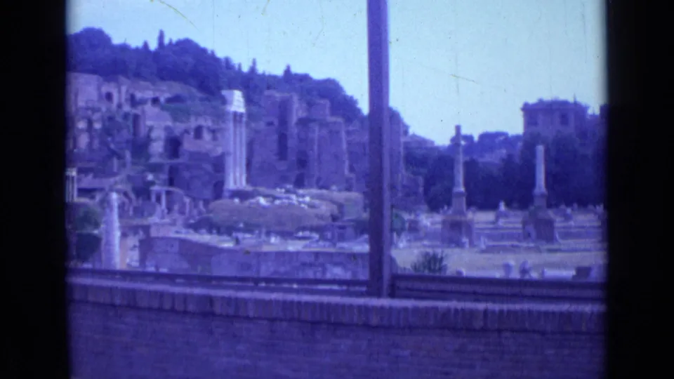 Visitor-shot Rome footage shows ancient columns and crumbling structures, with a distant building and trees beyond the ruins.