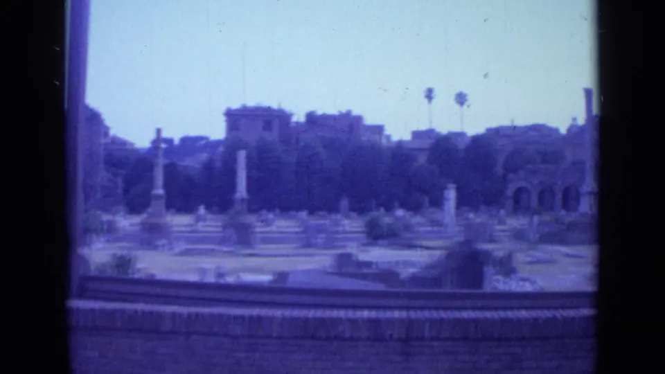 1962 Rome travel-film frame looking through a dark divider toward tomb markers, classical columns, ruins, trees, and hillside buildings.