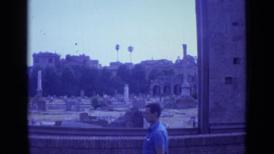 1962 Rome 8mm frame with a man walking in the foreground before cemetery grounds, columns, arched ruins, palms, and buildings.