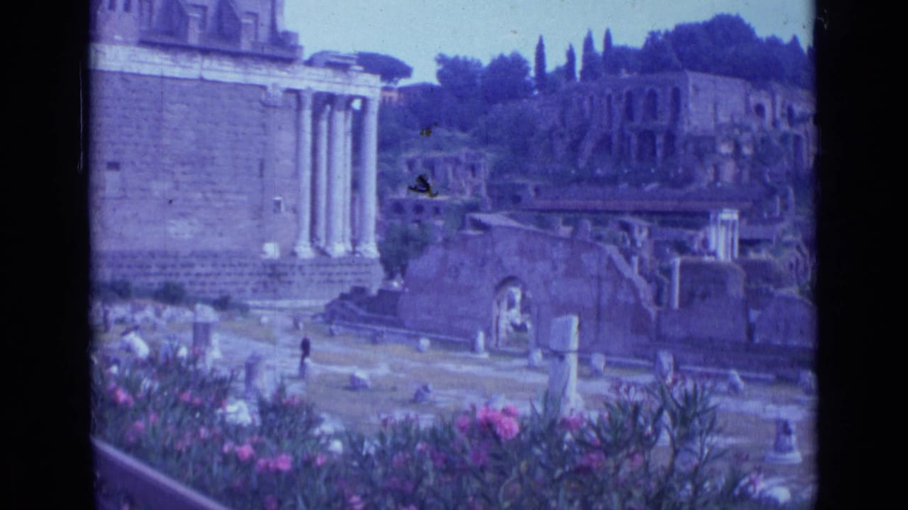 1962 Rome 8mm frame showing classical columns, weathered ruins, pink flowers, and distant visitors under bright daylight.