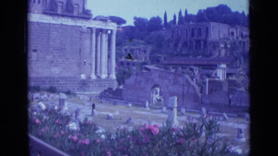 1962 Rome 8mm frame showing classical columns, weathered ruins, pink flowers, and distant visitors under bright daylight.
