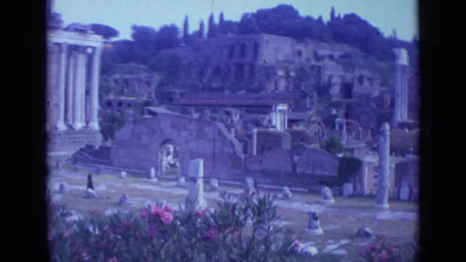 Rome 1962 8mm frame with crumbling masonry, upright columns, pink flowers, trees, and distant visitors crossing the ruins.