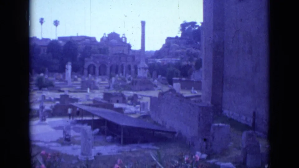1962 Rome travel-film frame showing a wider ruin field with columns, arched stonework, trees, visitors, and a wall edge at right.