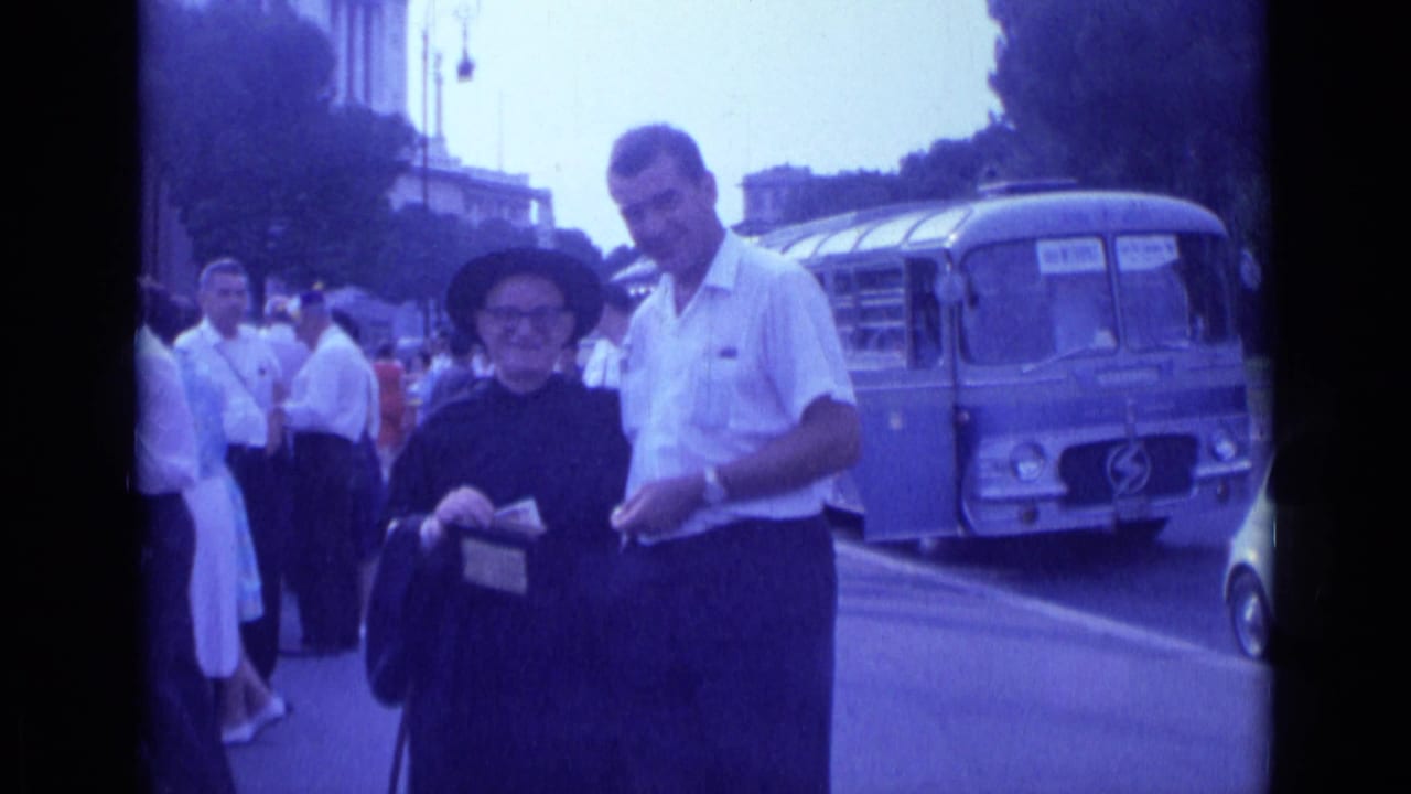 Man in a white shirt stands beside an older woman in dark clothing near a blue bus.