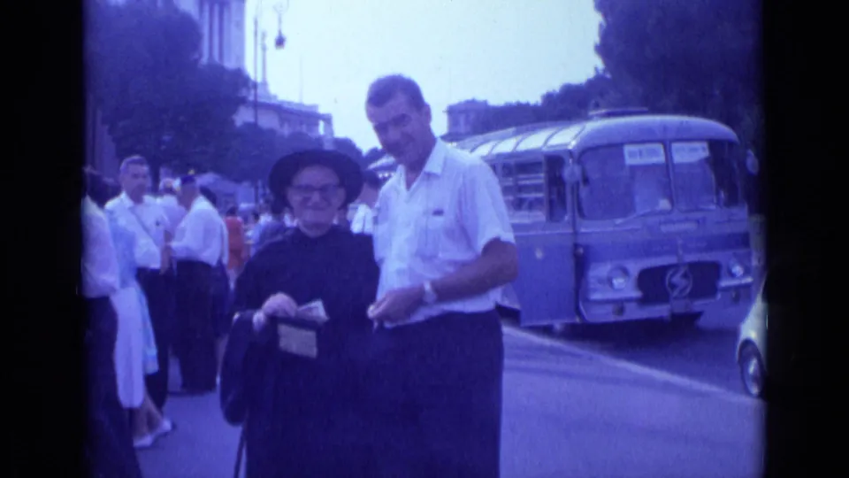 1962 Rome sidewalk scene with a man in a white shirt and older dark-clad woman looking down near a parked blue bus.