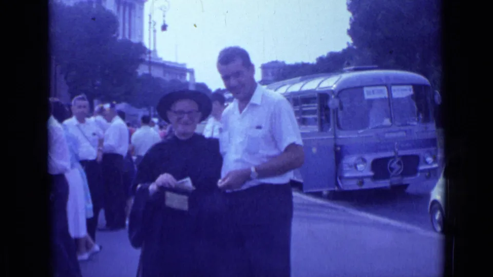 1962 Rome street frame showing an older woman holding an object while a man stands beside her near a blue bus.