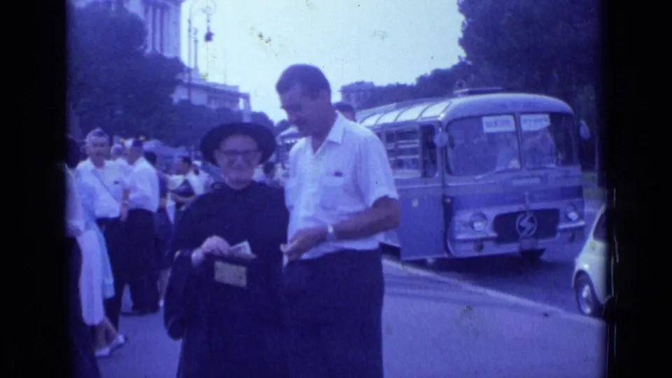 1962 Rome home-movie frame of a man and older woman by the roadway, with a bus, car and pedestrians behind them.