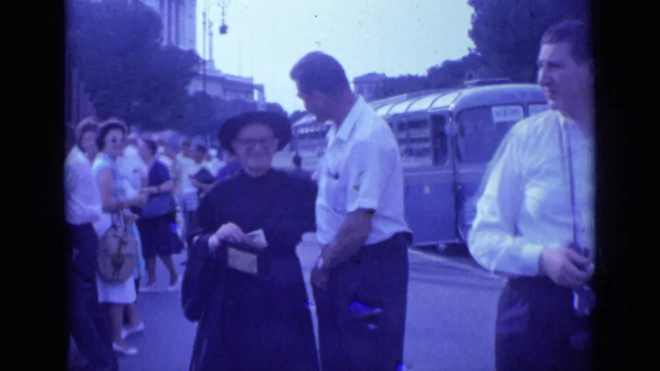 In a 1962 Rome street scene, a nun holds a donation box as a man stands close and pedestrians gather near a parked bus.