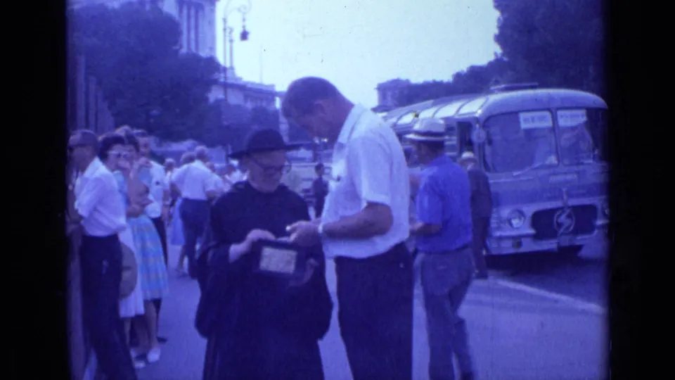 A 1962 Rome street moment shows the nun looking down at the donation box as the man stands close, with a bus and pedestrians in the background.