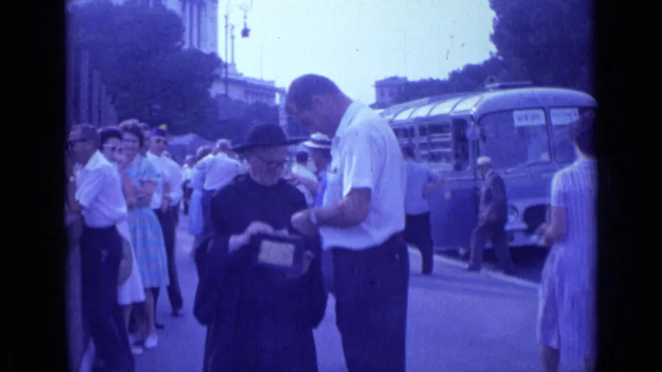 In 1962 Rome, a man in a white shirt appears to interact with a robed religious figure holding a small collection box near pedestrians and a bus.