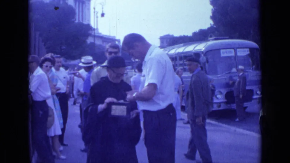 In the 1962 Rome home-movie frame, the donation exchange remains centered as a suited pedestrian and bus pass in the background.