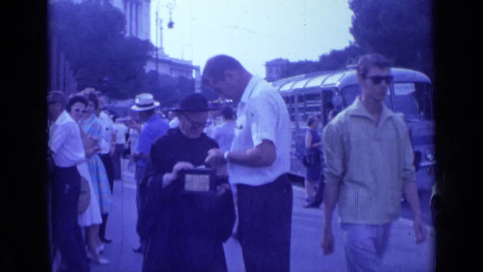 A passerby in sunglasses crosses the 1962 Rome street frame while the robed figure, white-shirted man, crowd, and bus remain visible.