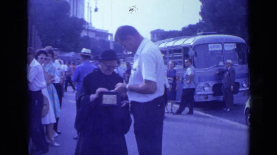 In 1962 Rome, a priest and a man focus on a small item while tourists and a blue bus fill the street background.
