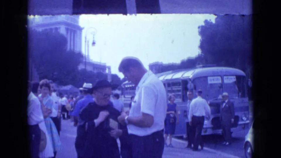 Rome home-movie frame from 1962 showing a priest and man exchanging attention over a small object as tourists and a bus remain nearby.