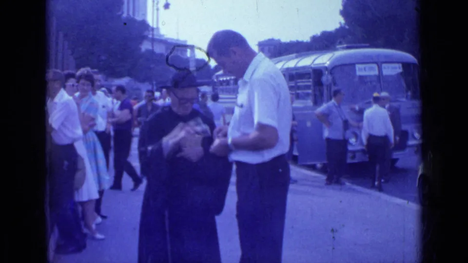 1962 Rome street footage shows the priest holding a small item while the man in white looks down and pedestrians pass near the bus.