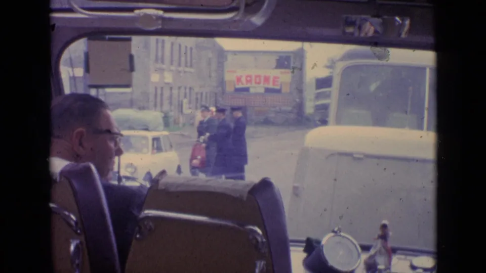 1962 Switzerland home-movie frame from inside a vehicle, with a man in glasses in the foreground and pedestrians and vehicles outside.