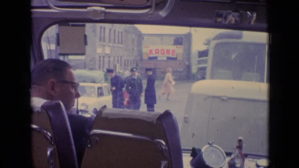 1962 Switzerland street scene seen through a vehicle windshield, with a seated man in foreground, police, pedestrians, cars, and a Krone sign.