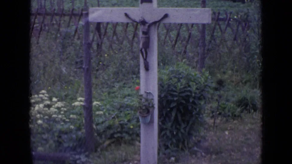 1962 Switzerland home-movie frame showing a wooden roadside crucifix with a figure of Christ, flowers, greenery, and a simple fence behind it.