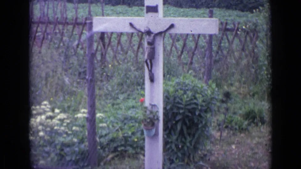 1962 Switzerland frame of a wooden roadside crucifix with a small plaque and religious figure, framed by flowers, greenery, and a rustic fence.