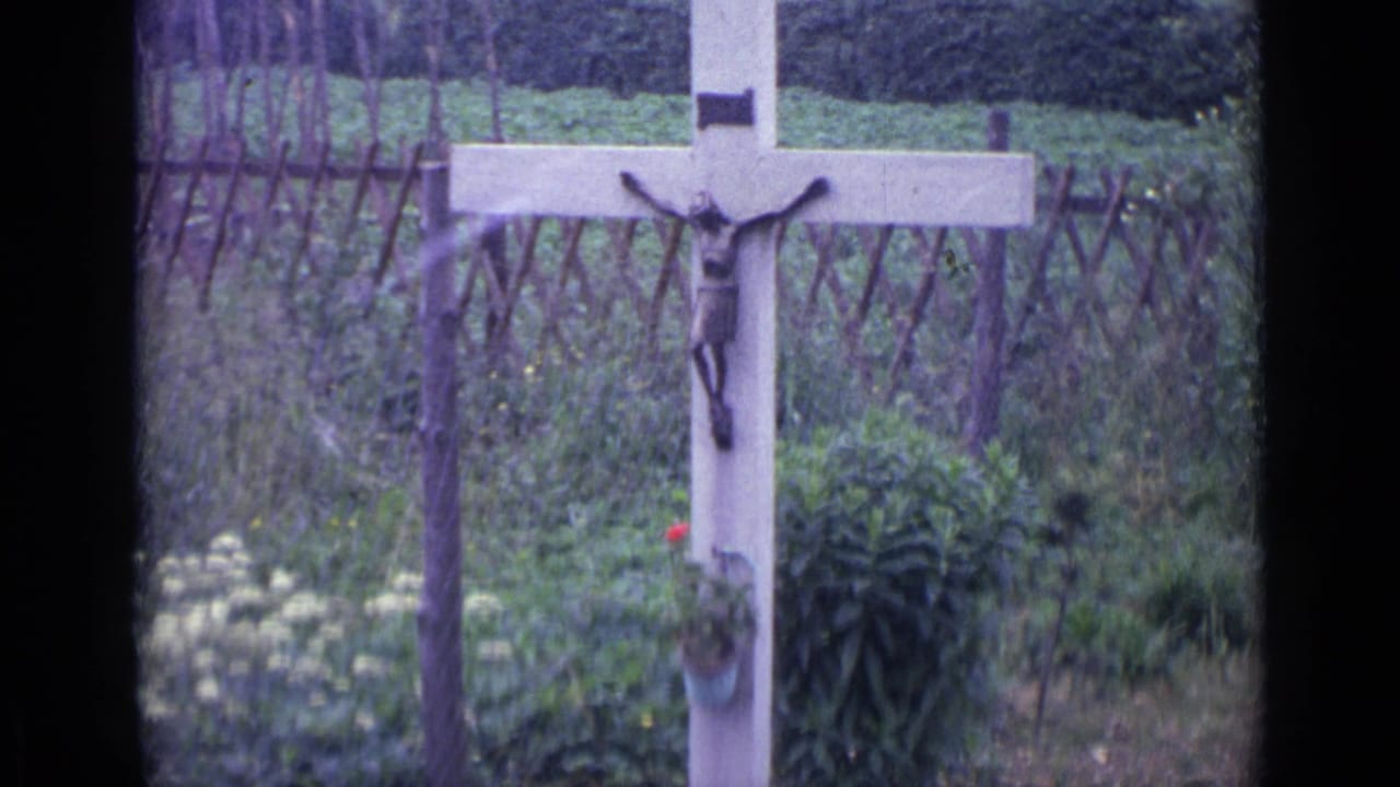 1962 Swiss home-movie frame showing a garden crucifix mounted on a pale cross, with plants and a rustic fence in the background.