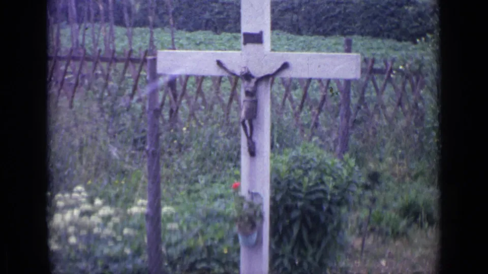 1962 Swiss home-movie frame showing a garden crucifix mounted on a pale cross, with plants and a rustic fence in the background.