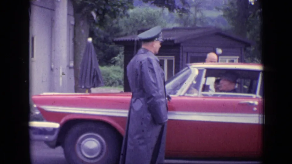 1962 Swiss home-movie frame showing a uniformed man in a cap beside a red classic car, with seated passengers partly visible.