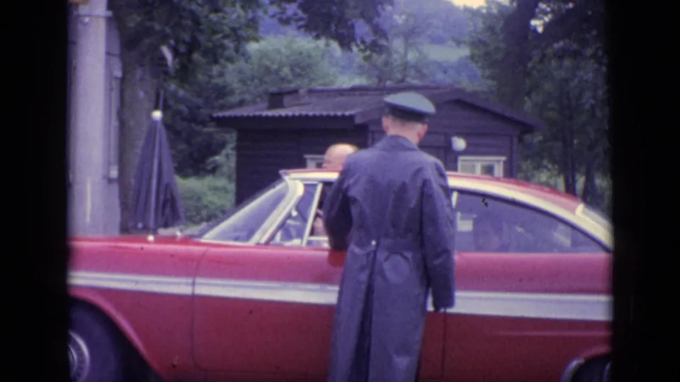 1962 Switzerland frame showing the back of a uniformed man beside a red classic car, with passengers and a small wooden structure behind.