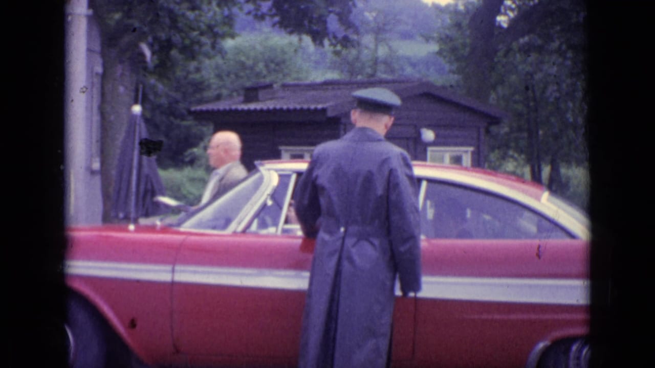 Police officer in a long coat stands beside a red classic car near trees.