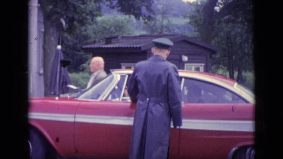 In 1962 Switzerland, a uniformed officer stands near a red classic car while a suited man is partly visible in a wooded roadside area.
