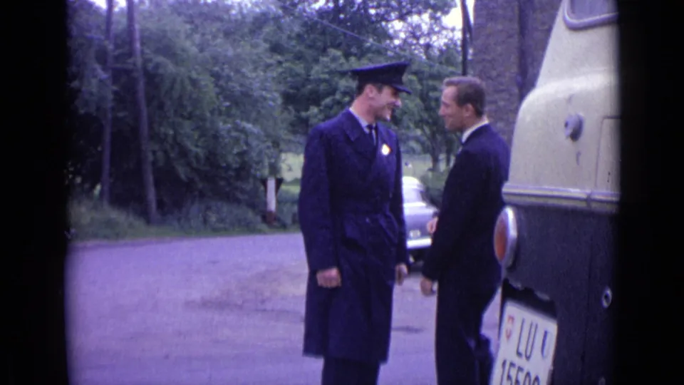A Swiss police officer in a dark coat and cap appears to speak with a suited civilian next to a cream vehicle or coach.