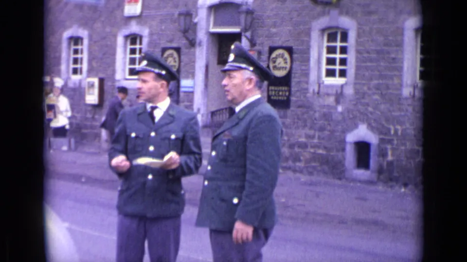 Two uniformed officers stand outside a stone building in Switzerland, with one holding papers or a notepad near posted signs.